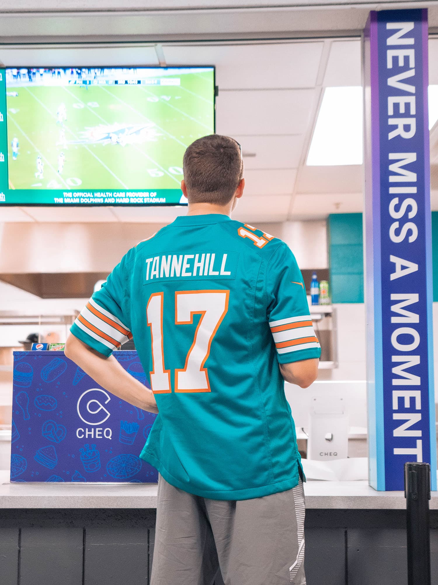 Miami Dolphin’s fan wearing a Tannehill jersey watching the game on a TV while waiting for his order.